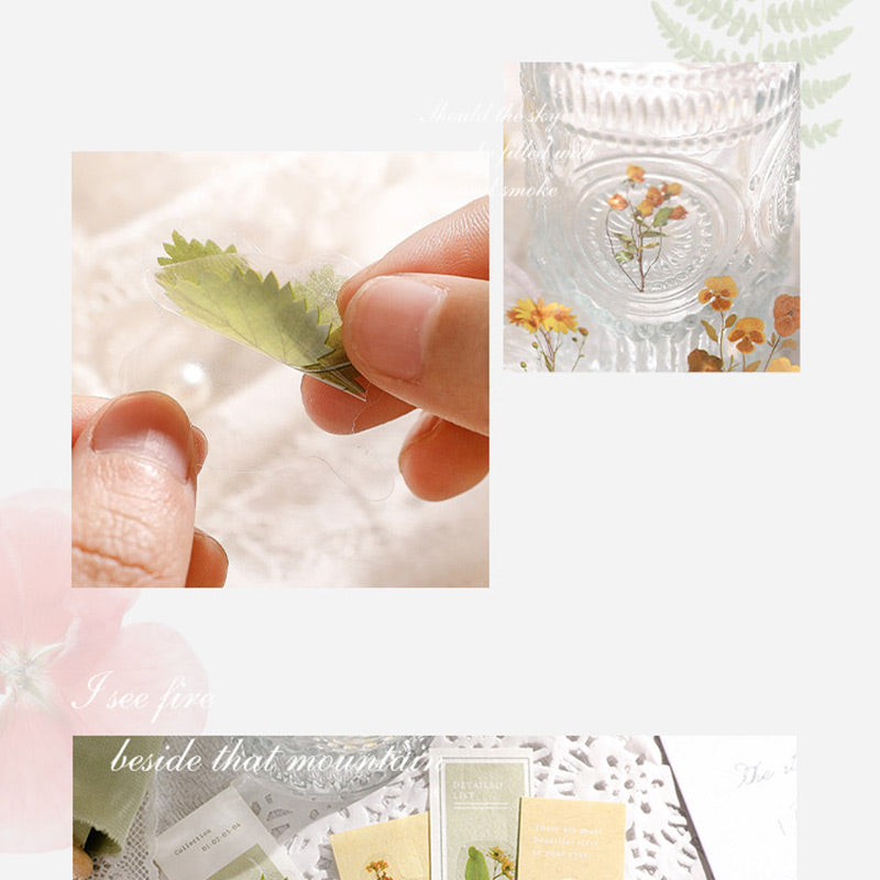 Fingers peeling a green leaf sticker from its backing, with a glass jar of stickers and bookmark cards on lace