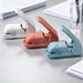 White, coral pink and light blue stapleless staplers on a grey desk beside loose paper and a pen holder