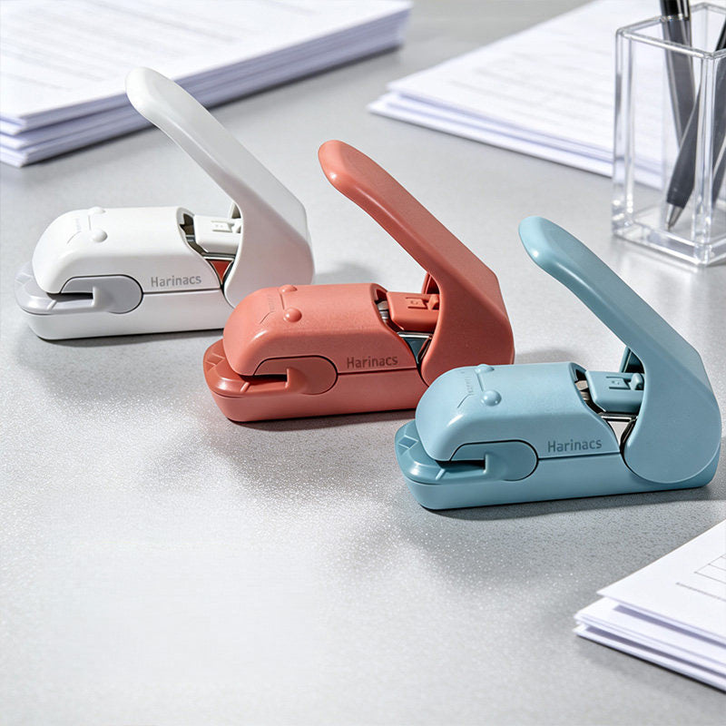 White, coral pink and light blue stapleless staplers on a grey desk beside loose paper and a pen holder