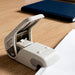 Open pale white stapleless stapler on a wooden desk beside a sheet of paper, with a notebook in the background
