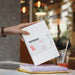 Hand lifting white Paperideas 2027 unbound A5 planner with belly band from a stack beside an iced drink on a cafe table