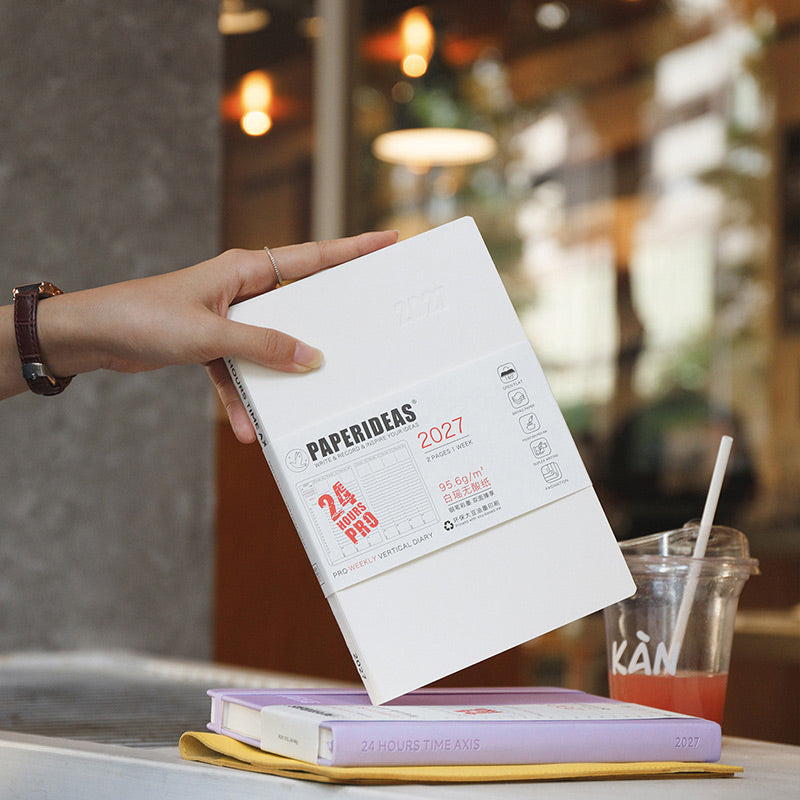 Hand lifting white Paperideas 2027 unbound A5 planner with belly band from a stack beside an iced drink on a cafe table