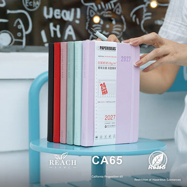 Six Paperideas 2027 A5 hardcover planners in black, red, pink, smoke blue, mint and lilac upright on a blue cafe stool