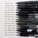 Fifteen black 0.5mm gel and rollerball pens lined up beside handwriting samples on grid paper to compare line width
