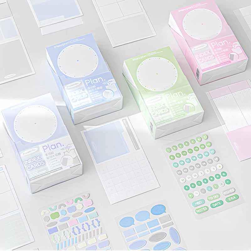 Blue, green and pink index card boxes with sticker sheets and blank planner cards scattered on a light desk