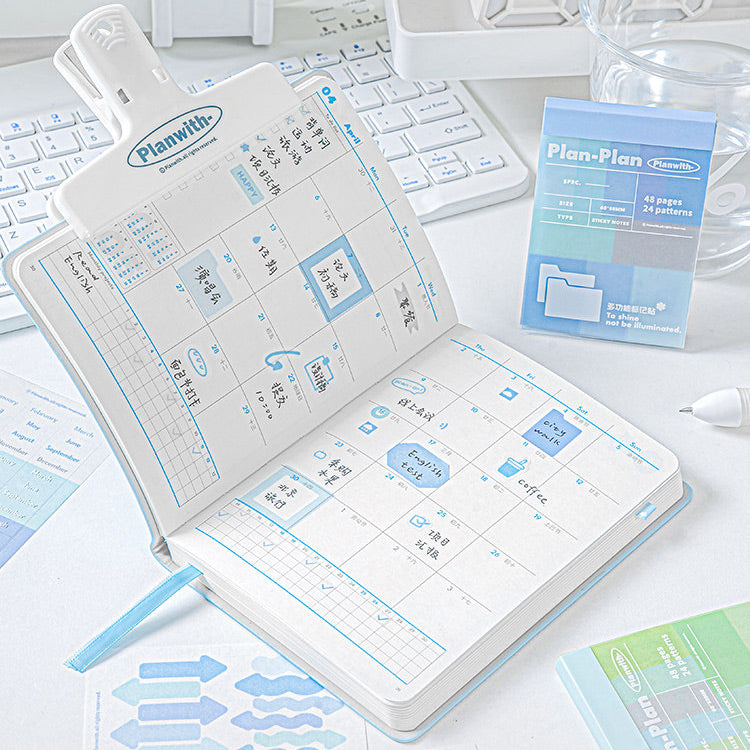 An open planner with a weekly calendar layout and blue sticky notes, shown on a white desk with a keyboard, glass, and a small blue package labeled Plan-Plan.