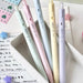 Five pastel C-JJ20 retractable gel pens standing in a white desk tray beside lined notes