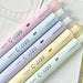 Five pastel C-JJ20 gel pens in pink, blue, purple, yellow and green resting on lined notebook paper