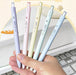 Hand holding five pastel purple, pink, yellow, green and blue C-JJ20 gel pens above a laptop keyboard